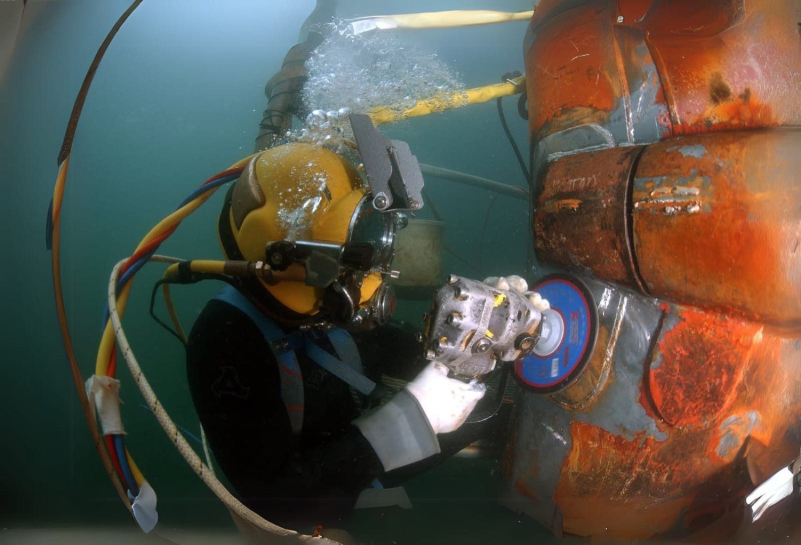 Surface-supplied diver in a yellow helmet running a grinder against a corroded steel pile underwater