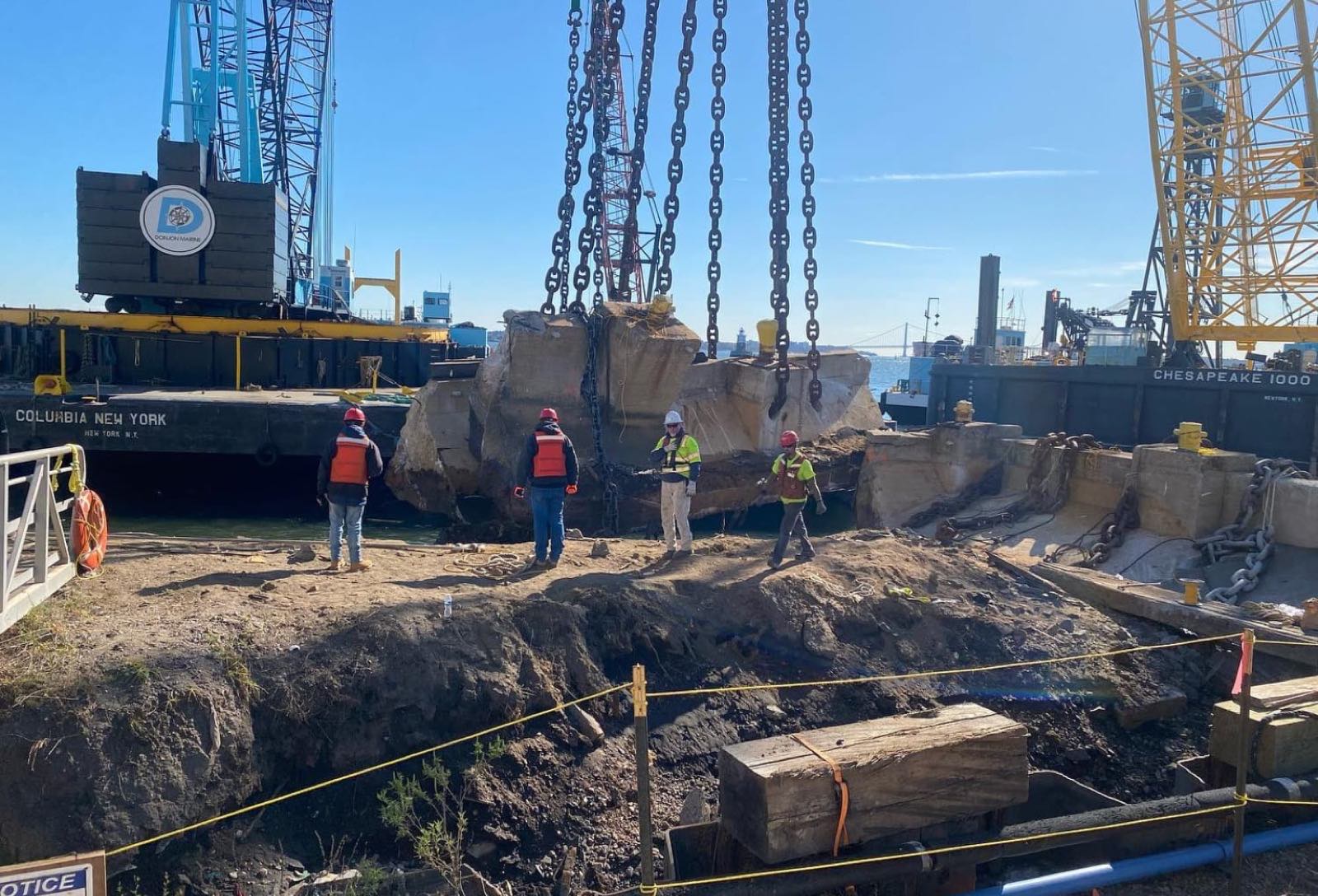Crane lifting broken concrete sections on chains between two barges while four crew members in hard hats stand on the bank